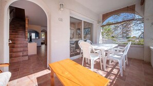 A sunlit patio with a wooden table and white plastic chairs, a staircase leading to a kitchen, and a large window with a view of trees. - R141 - C6 Manila Resort Patio (Calafell)