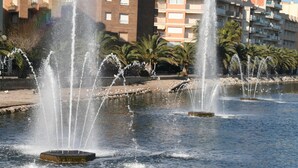 A fountain with multiple jets of water in a public square, surrounded by palm trees and buildings. - R141 - C6 Manila Resort Patio (Calafell)