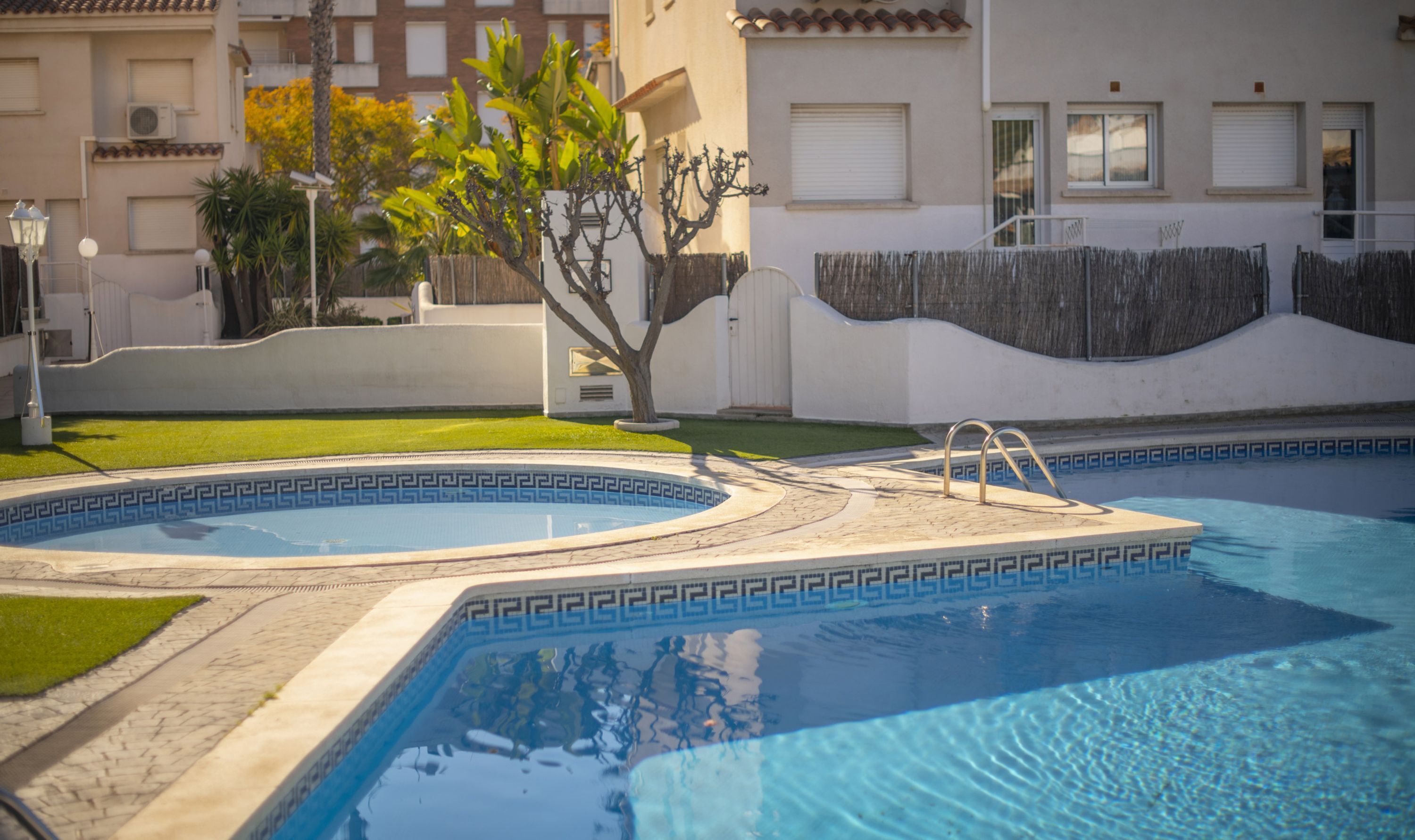 A swimming pool with a geometric tile pattern, surrounded by a paved area and a small tree.