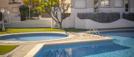 A swimming pool with a geometric tile pattern, surrounded by a paved area and a small tree.