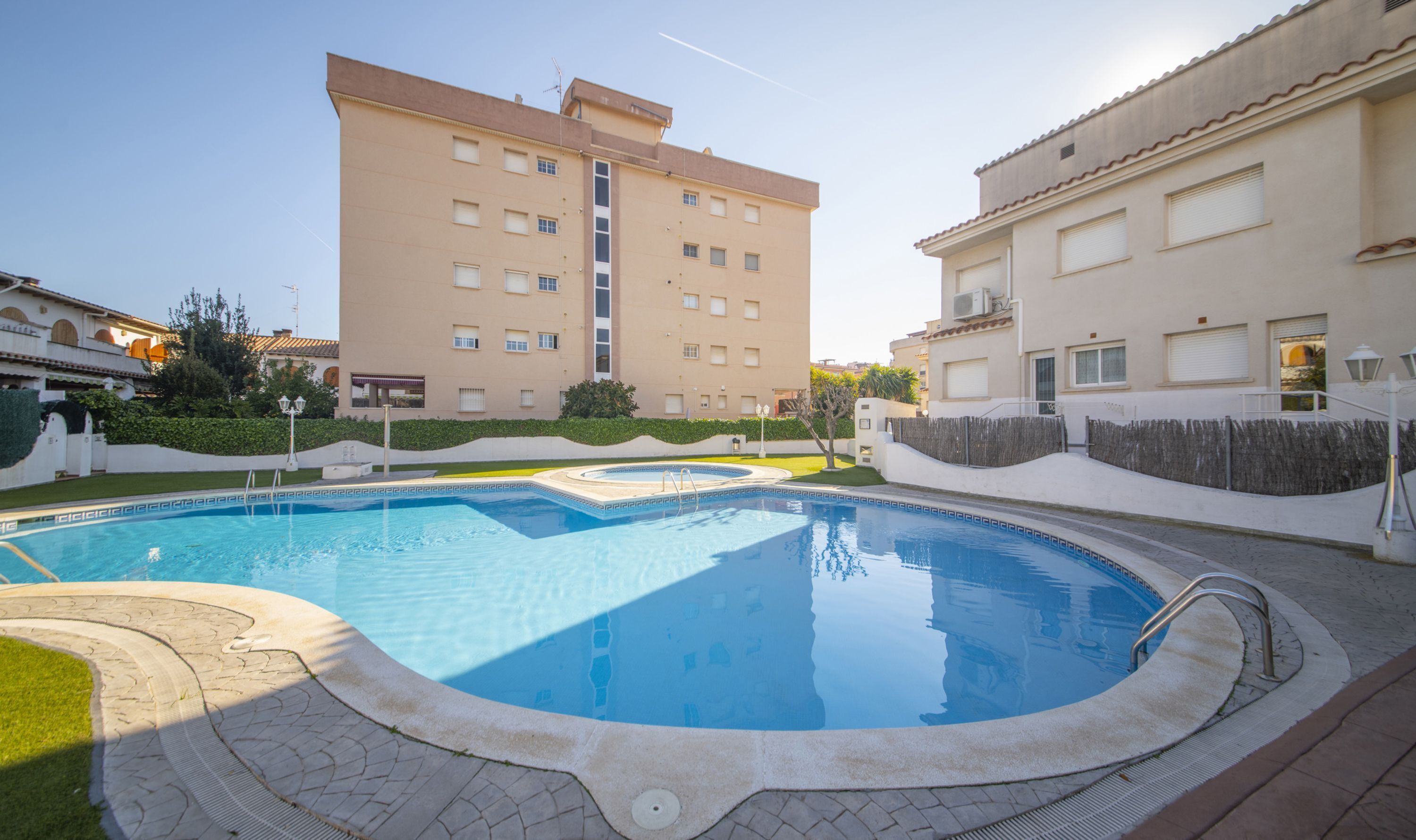 Une piscine avec une terrasse carrelée, entourée d’un petit muret et bordée, au loin, d’un grand immeuble.
