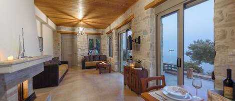 A spacious living area with a fireplace, stone walls, wooden ceiling, and large windows offering a view of the outdoors.