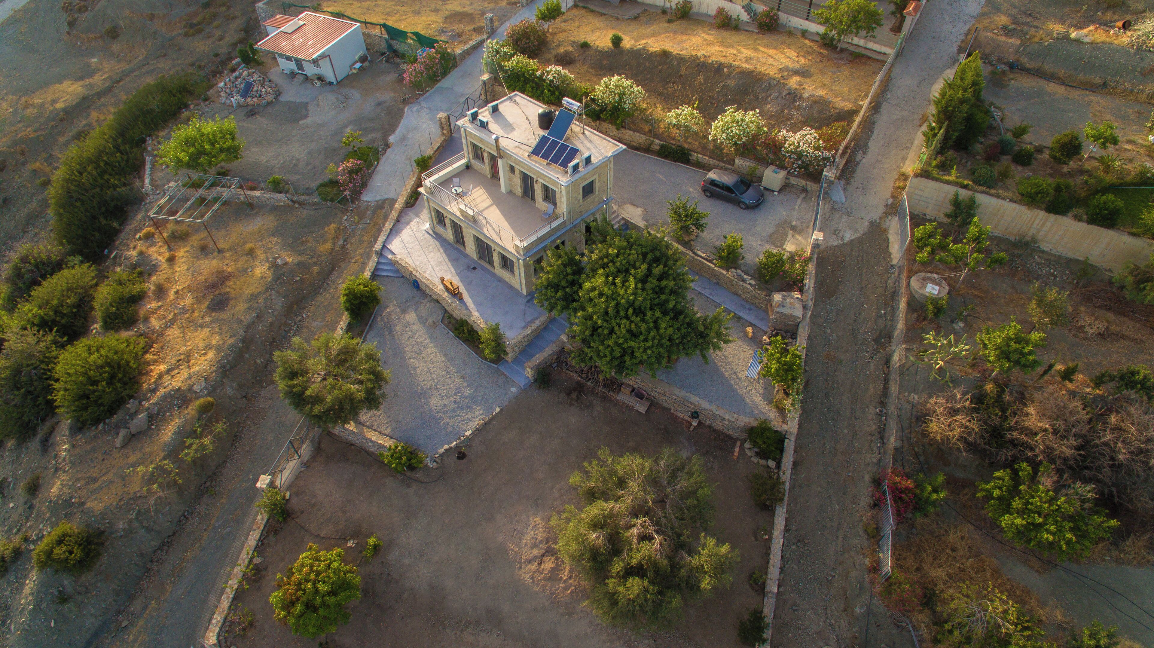An aerial view of a two-story house with a solar panel on the roof, surrounded by trees and a small garden.