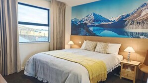 Ein Hotelzimmer mit einem großen Bett, zwei Nachttischen mit Lampen, einem Fenster mit Vorhängen und einem Wandbild von einer Berglandschaft.