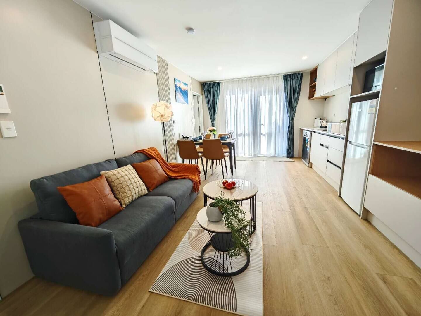 A modern living room with a grey sofa, a dining table with chairs, and a kitchen area with white cabinets and a refrigerator.