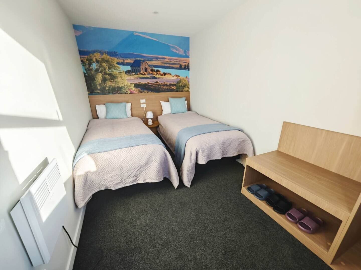 A small, well-lit room with two beds, a wooden nightstand, a shelf with shoes, and a mural of a landscape.
