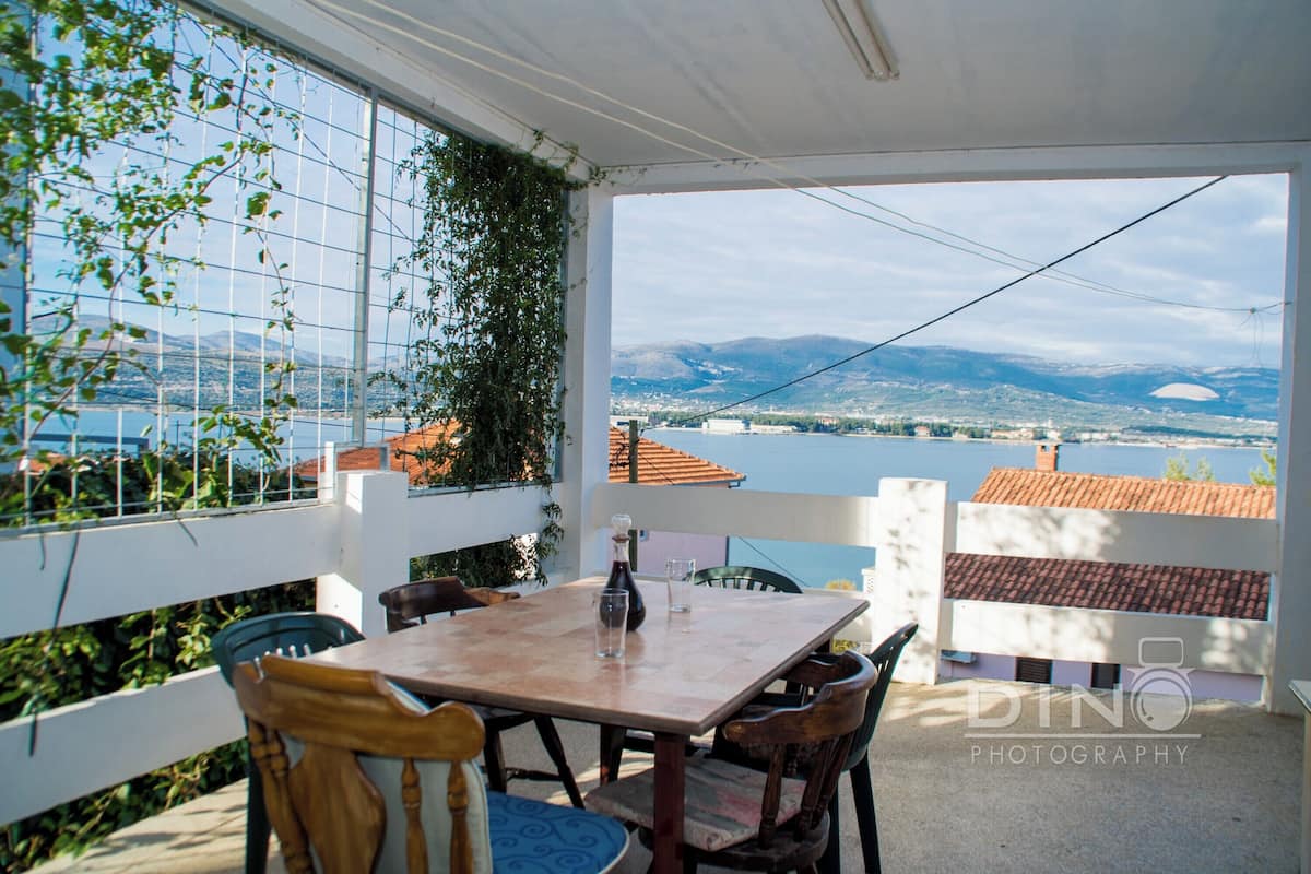 A terrace with a table and chairs, a view of a lake and mountains, and a white building with a red roof.