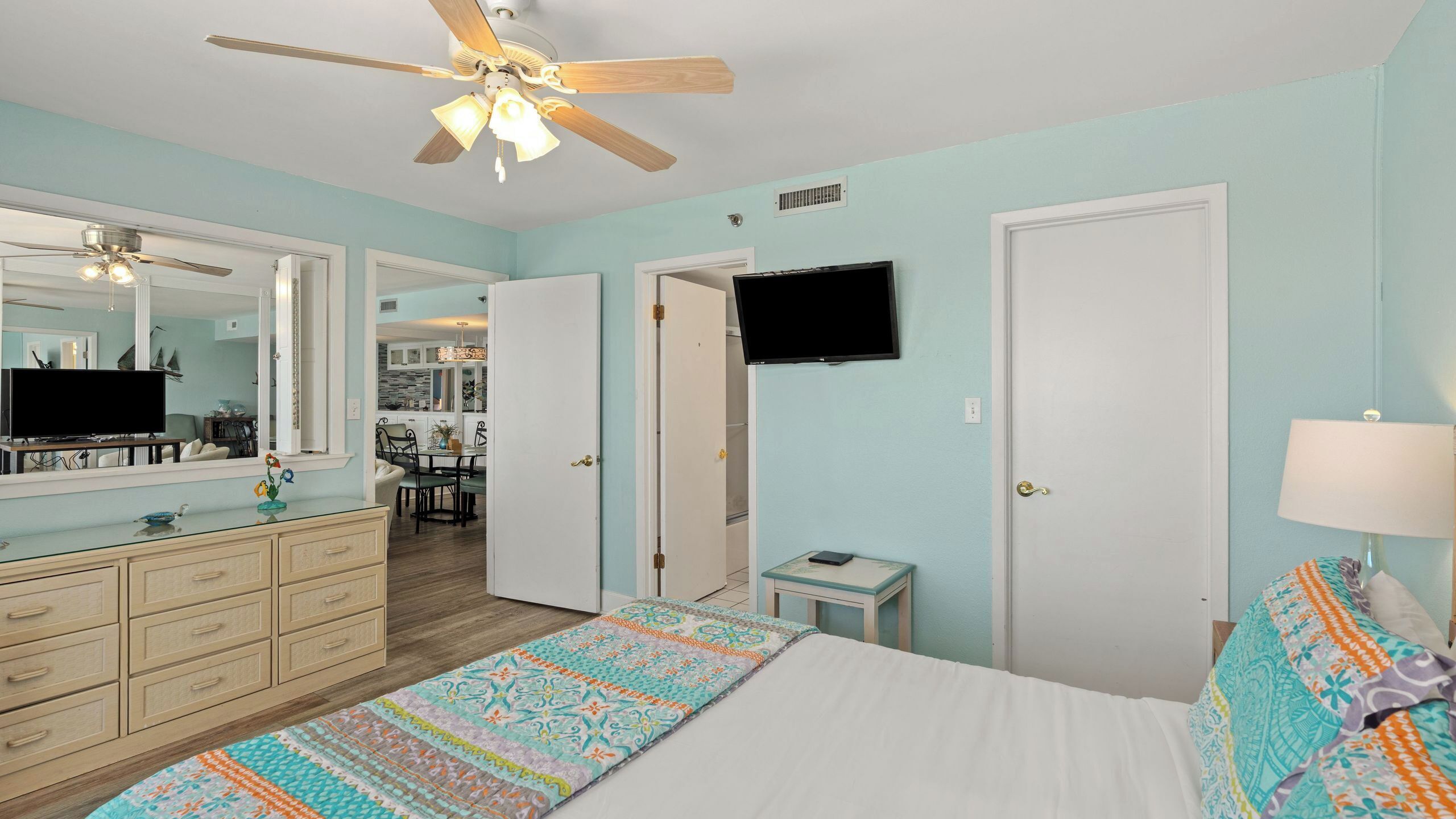 A bedroom with a bed, dresser, TV, ceiling fan, and a small table with a lamp.