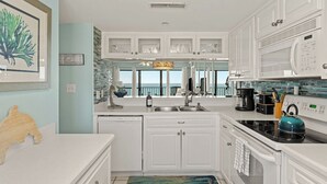 A modern kitchen with white cabinetry, a coffee maker, a kettle, and a view of the ocean. - Gull Watch by Avantstay Beachfront Condo (Panama City Beach)