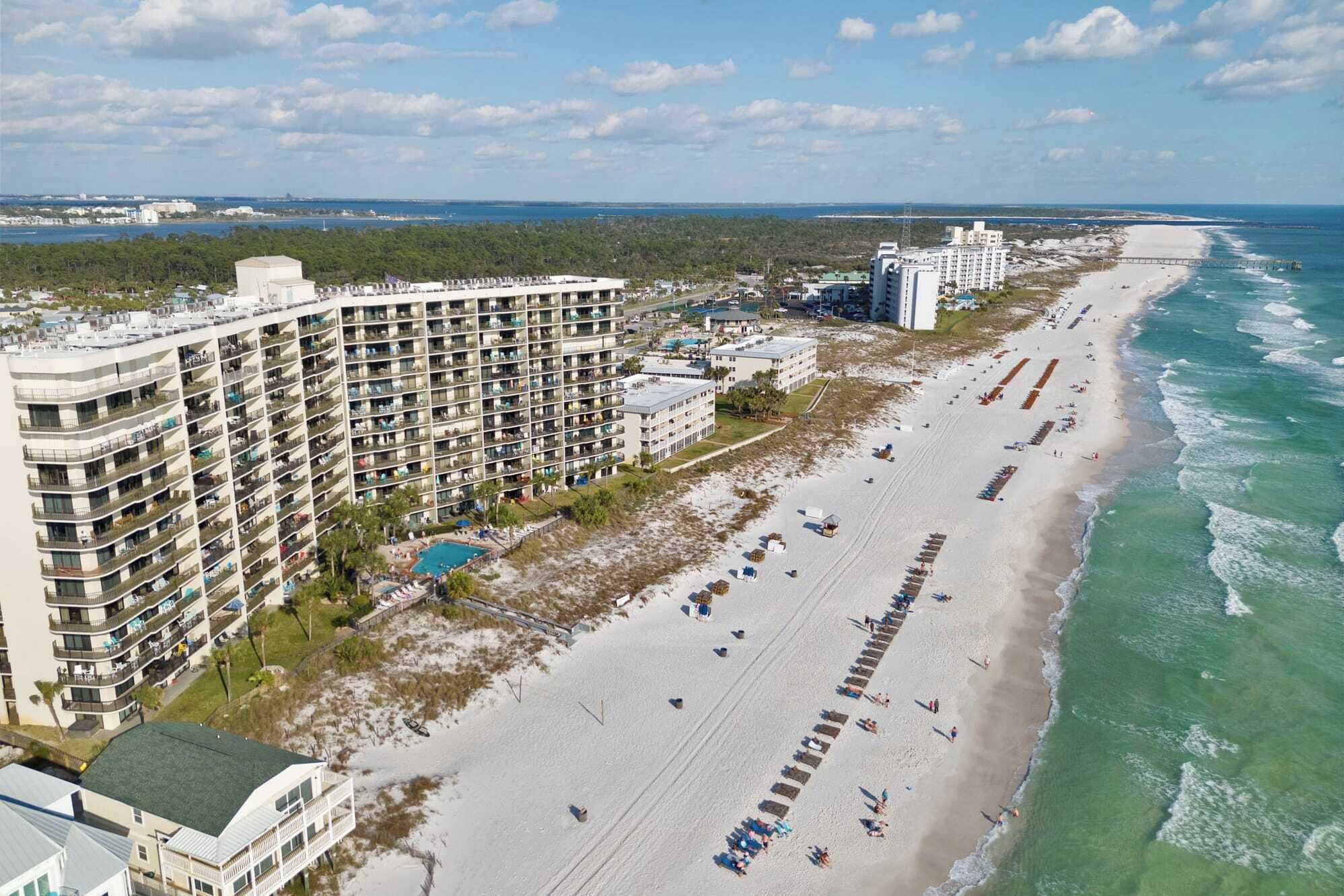 A high-rise hotel with multiple balconies, a beach with rows of umbrellas, and a clear view of the ocean.