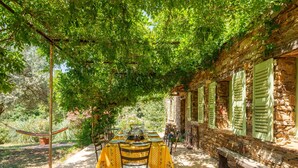 Un área de comedor al aire libre con techo, con una mesa dispuesta para dos personas, rodeada de muros de piedra y contraventanas verdes.