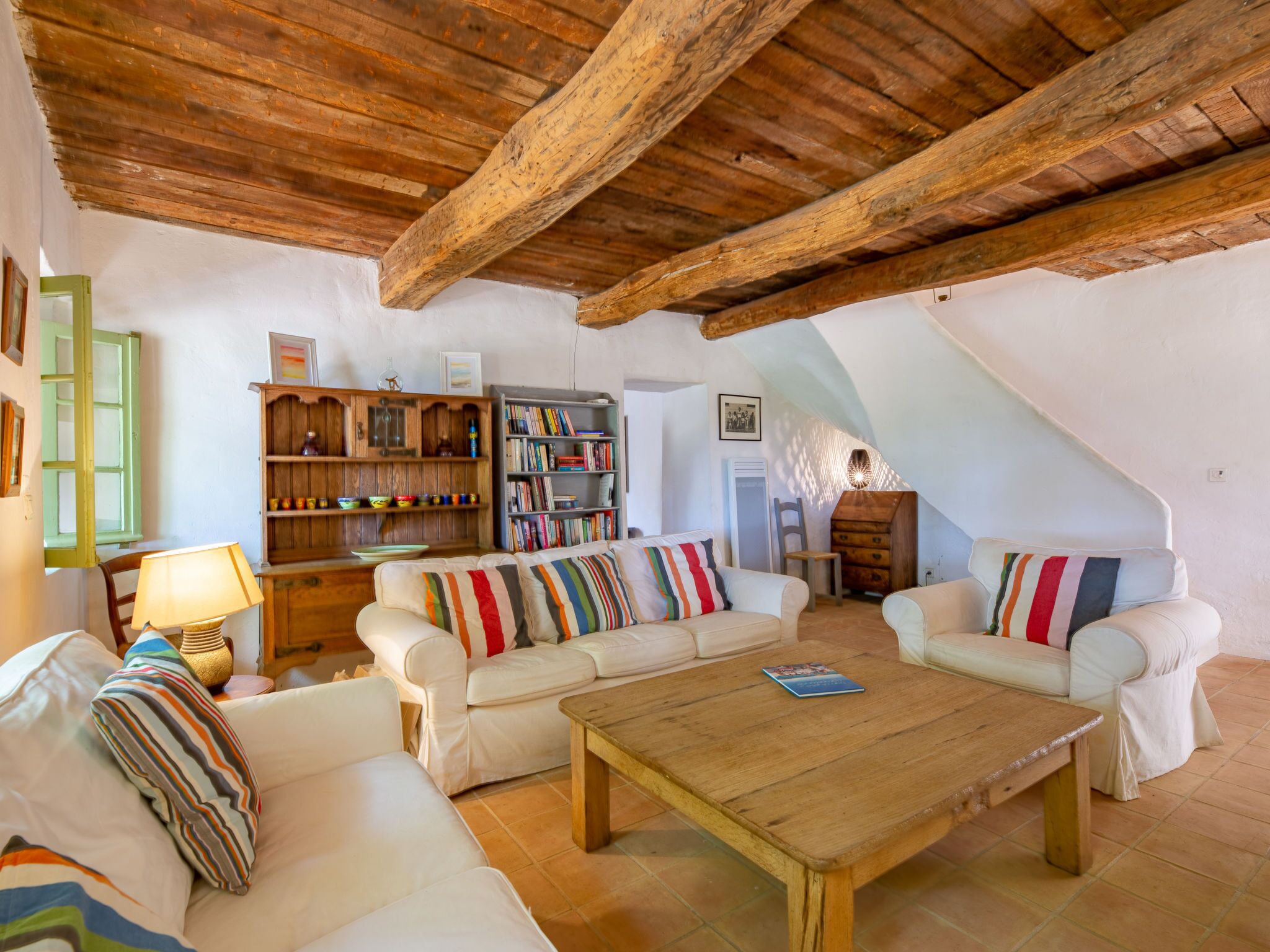 A cozy living room with a wooden ceiling, white sofas, a coffee table, and a bookshelf filled with books and decorative items.