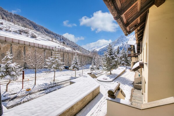 A snow-covered courtyard with a wooden building, a stone wall, and a mountain backdrop.