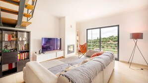 Un salon moderne avec un canapé, un téléviseur, une bibliothèque et une vue sur la verdure grâce à de grandes fenêtres.