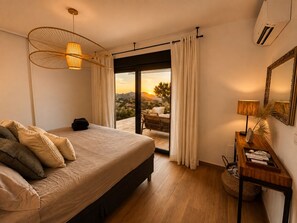 A bedroom with a bed, a nightstand, a lamp, a mirror, and a view of the outdoors.