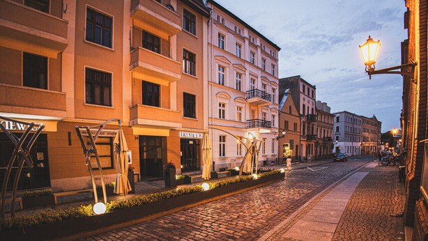 A cobblestone street lined with multi-story buildings, some with visible signage. - 2HomeRent Estrella 10 | Old Town | Spacious (Torun)