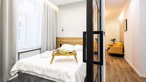 A hotel room with a bed, a wooden bedside table with a tray, a window with curtains, a wooden floor, and a yellow sofa in the adjacent living area. - 2HomeRent Estrella 10 | Old Town | Spacious (Torun)