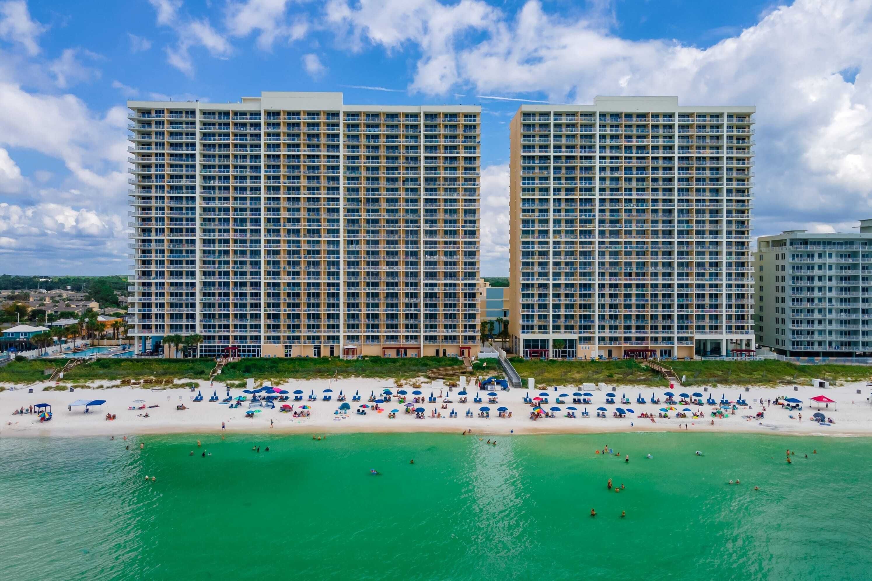 Een strandhotel met meerdere hoge gebouwen, een zandstrand met parasols en helder turquoise water.