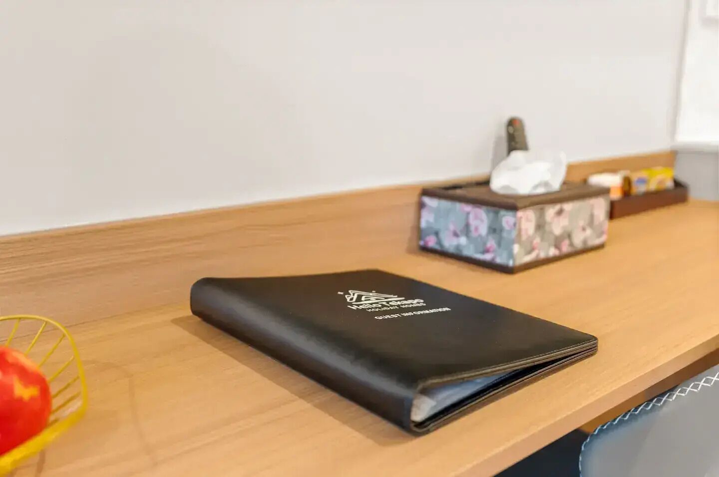 A neatly arranged hotel room with a wooden side table, a black guest information folder, a floral tissue box, and a yellow basket.