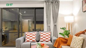 A modern living room with a grey sofa, checkered cushions, a small round table with a flower vase, a floor lamp, and a large window with curtains.