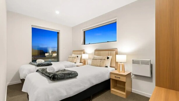 A hotel room with two beds, a nightstand with a lamp, a heater, and a view of the cityscape through the windows.