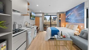 A modern hotel room with a large bed, a sofa, a small table with a bottle and glass, a TV, a kitchen area with a washing machine, and a large window with a view.