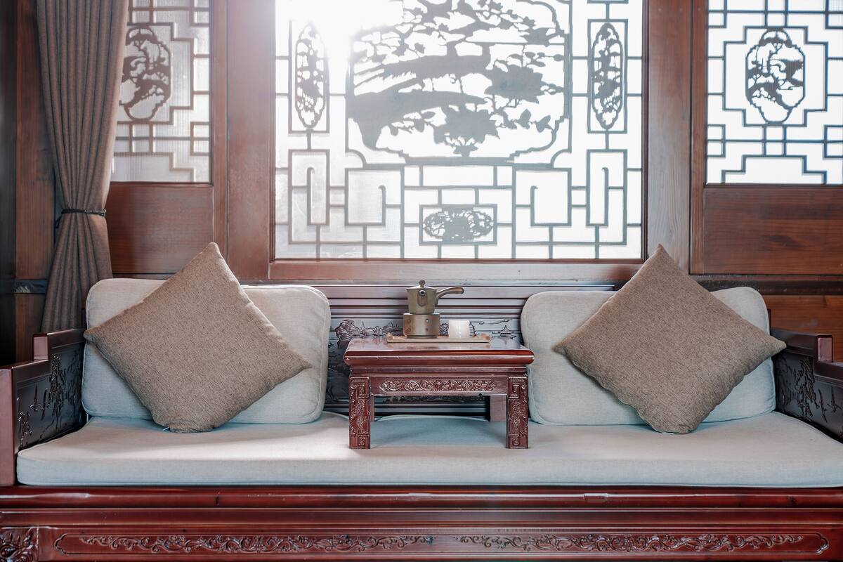 A traditional-style sofa with two pillows, a small wooden table with a teapot, and a decorative window with intricate patterns.