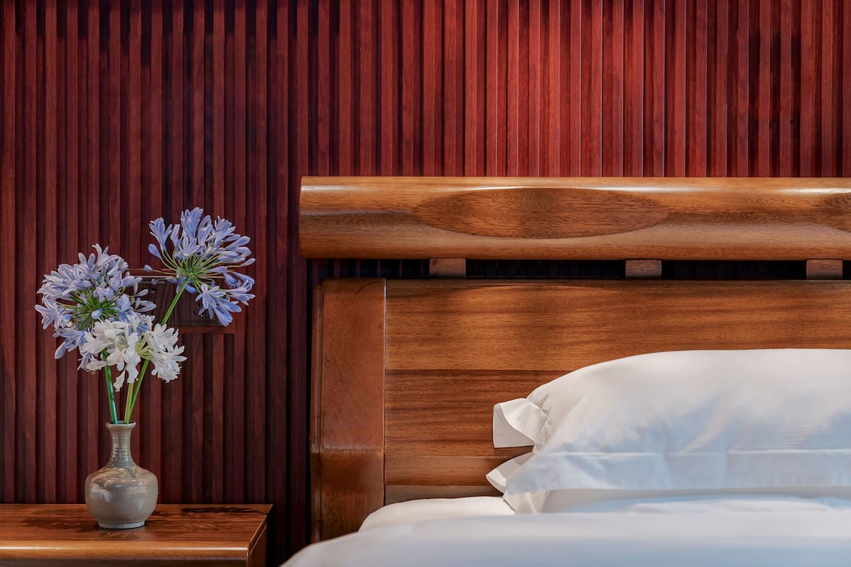 A neatly made bed with white linens, a wooden headboard, and a vase with flowers on a bedside table against a wooden wall.