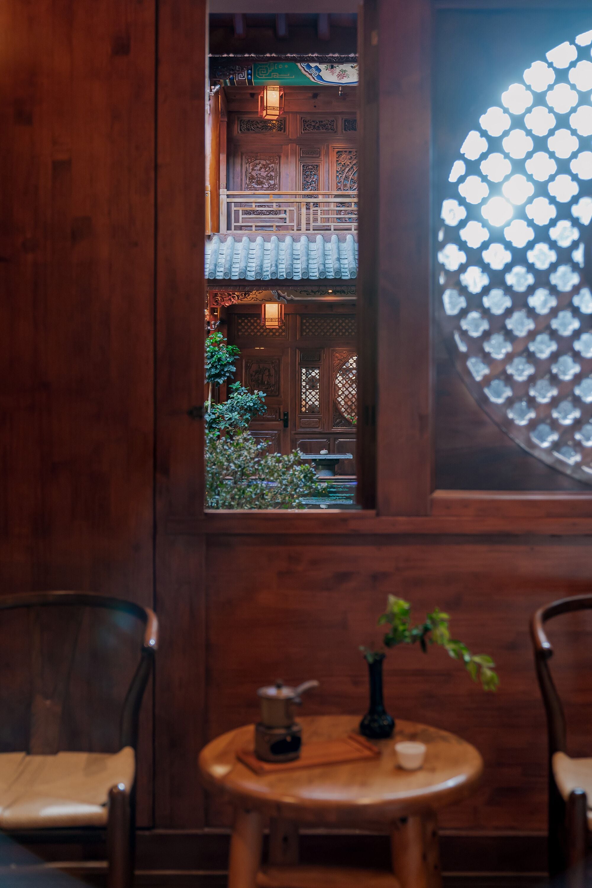 A wooden interior with a traditional Chinese-style window, a small round table with a plant, and two chairs.
