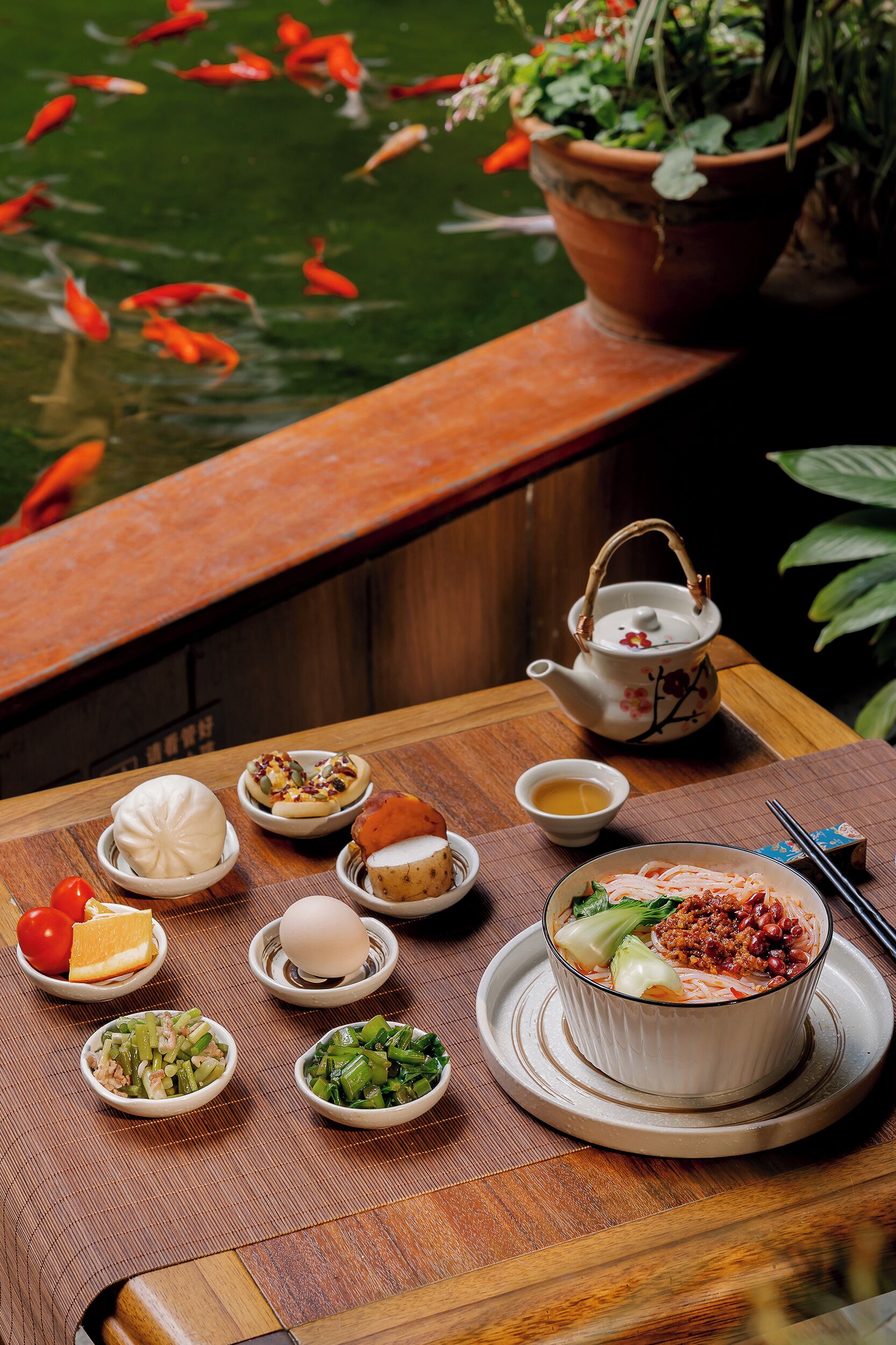 A table with various dishes, a teapot, and a view of a pond with koi fish.