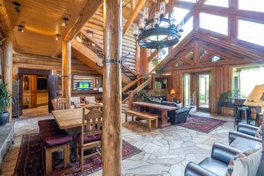 A spacious lodge interior with a stone floor, wooden furniture, a fireplace, and a staircase leading to upper levels.