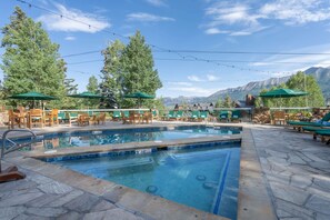 Zona de piscina con sillones y sombrillas, rodeada de árboles y montañas al fondo.
