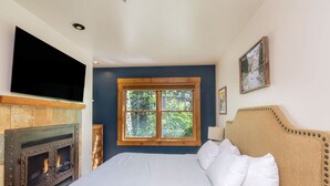 Een slaapkamer met een bed, een open haard, een televisie en een raam met uitzicht op bomen.