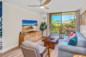 A living room with a sofa, a coffee table, a TV, and a balcony with a view of the ocean. - Hip Hawaiian Oceanfront Vistas (Kihei)