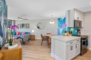 A modern kitchen with a built-in oven, a dining area with a round table and chairs, and a living room with a sofa and a ceiling fan.