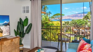 A living room with a large TV, a wooden cabinet, a plant, a sofa with colorful cushions, and a balcony with a view of the ocean and tropical plants. - Hip Hawaiian Oceanfront Vistas (Kihei)