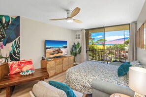 Een slaapkamer met een bed, een televisie, een plafondventilator en een balkon met uitzicht op het strand.