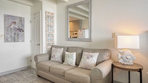 Un salón con un sofá color crema, tres cojines estampados, una mesita redonda con un pez decorativo y un espejo grande en la pared.
