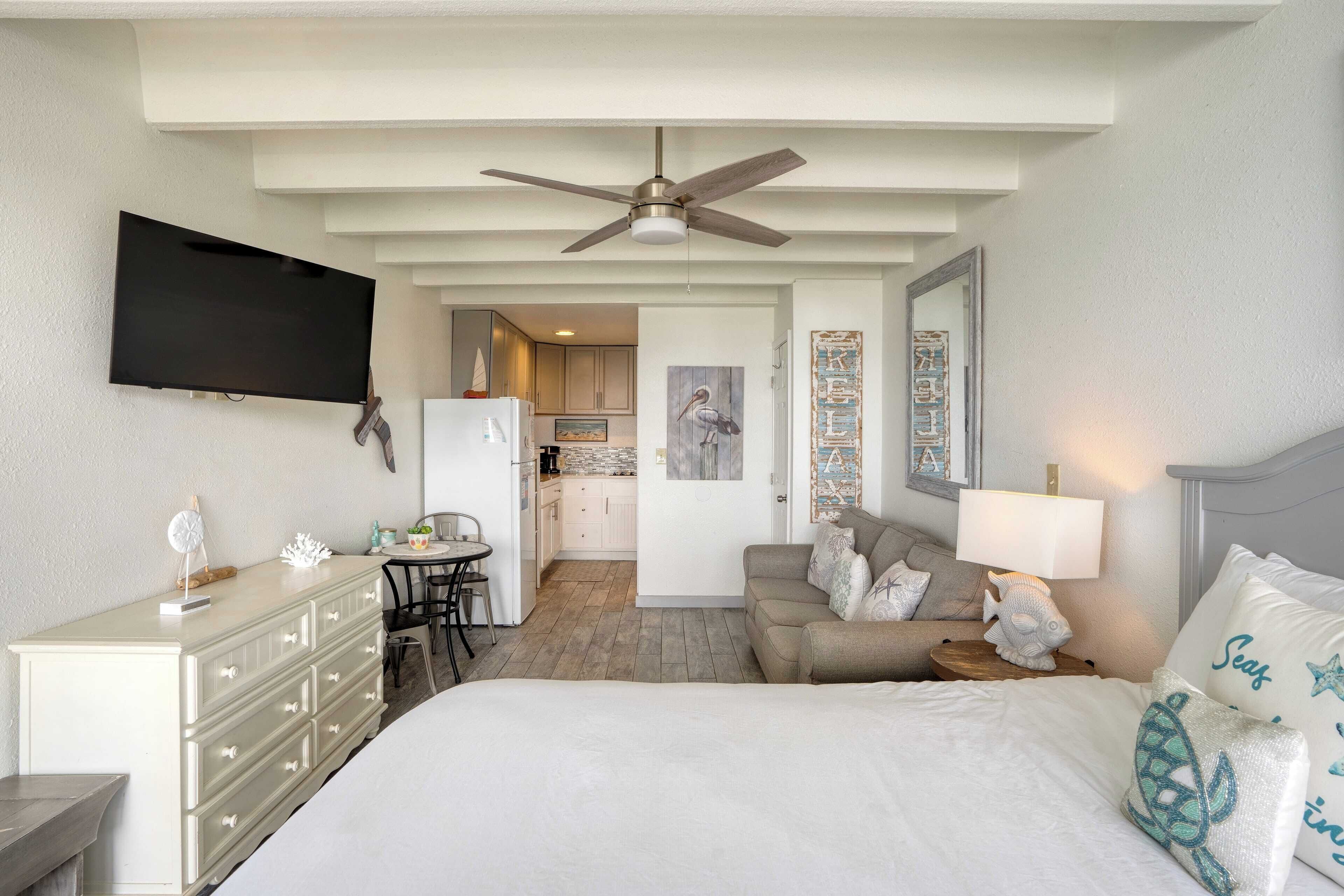 A bedroom with a bed, dresser, TV, ceiling fan, and a view into the kitchen.