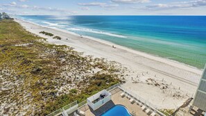 A beach view with a pool area, a pier in the background, and a building with a balcony overlooking the beach.