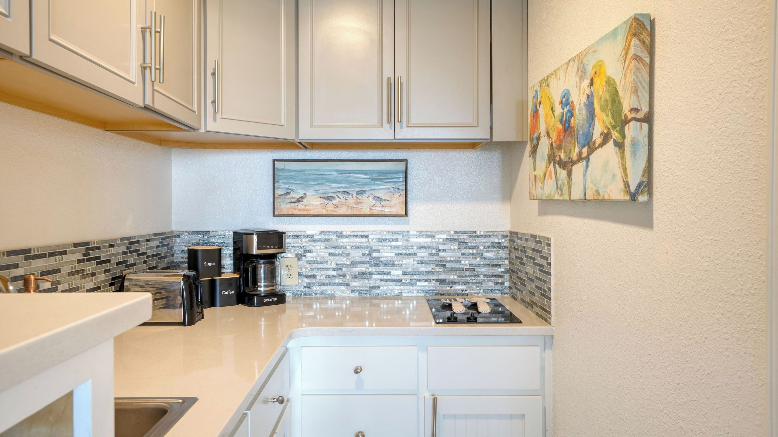A compact kitchen with white cabinets, a coffee maker, sugar and coffee containers, a tiled backsplash, and a painting of birds on the wall.
