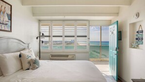 Un dormitorio con cama, ventana con contraventanas, puerta y vista a la playa.