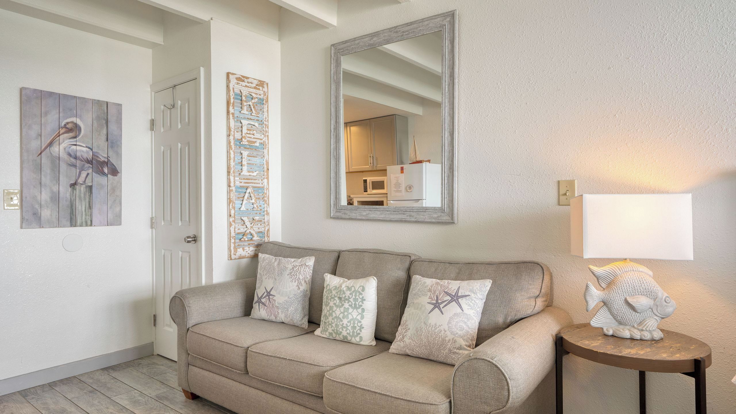 A neutral-toned living room with a beige sofa, decorative pillows, a large mirror, and a wall-mounted lamp.