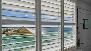 Una habitación con vista al océano y un paisaje urbano lejano, visibles a través de persianas blancas.