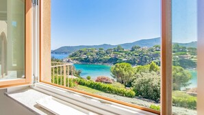 Ein Balkon mit Blick auf eine Küstenlandschaft, darunter ein klares blaues Meer, grüne Hügel und verstreute Gebäude.
