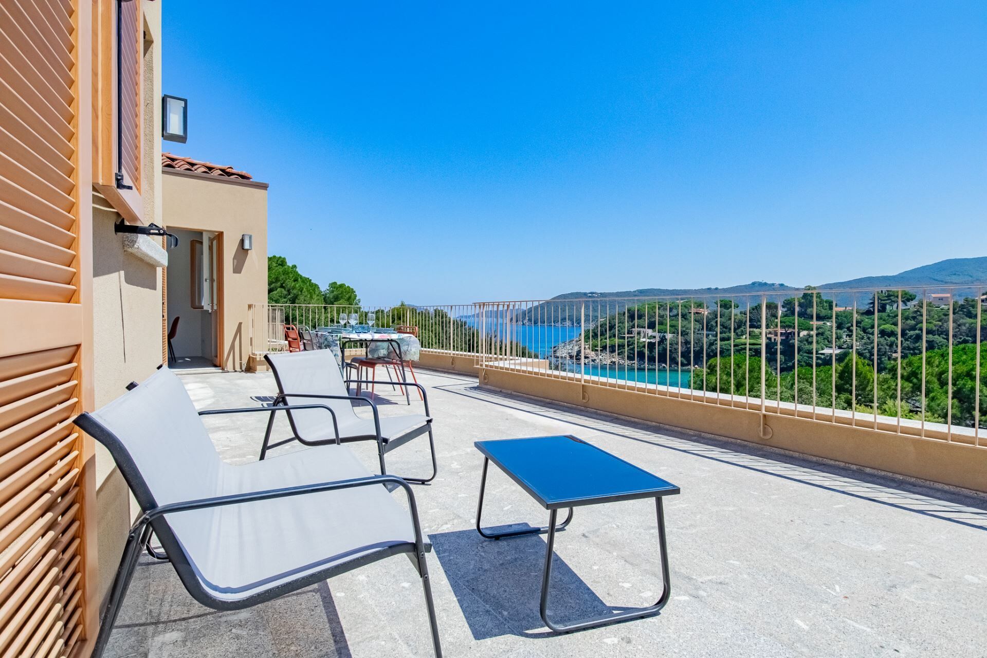 Een balkon buiten met een tafel en stoelen, met uitzicht op een schilderachtig kustlandschap met bergen en groen.