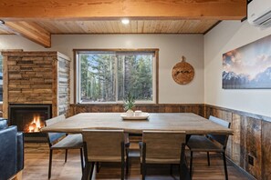 Ein Holzespeisetisch mit sechs Stühlen, ein Steinkamin, ein großes Fenster mit Blick auf Bäume und eine Wanduhr.