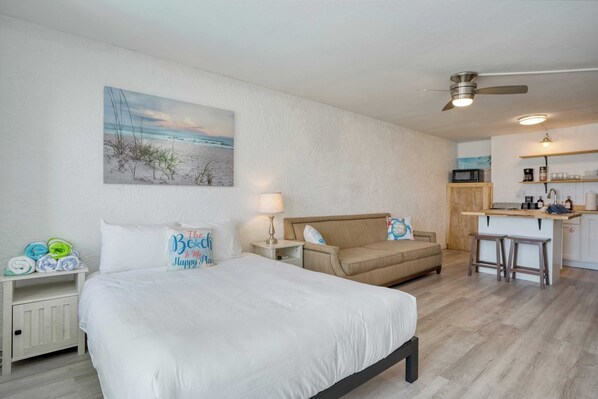 A bedroom with a bed, a nightstand, a bedside table, a lamp, a wall-mounted painting, and a kitchenette in the background. - Sun-kissed by Avantstay Beachfront Access (Panama City Beach)
