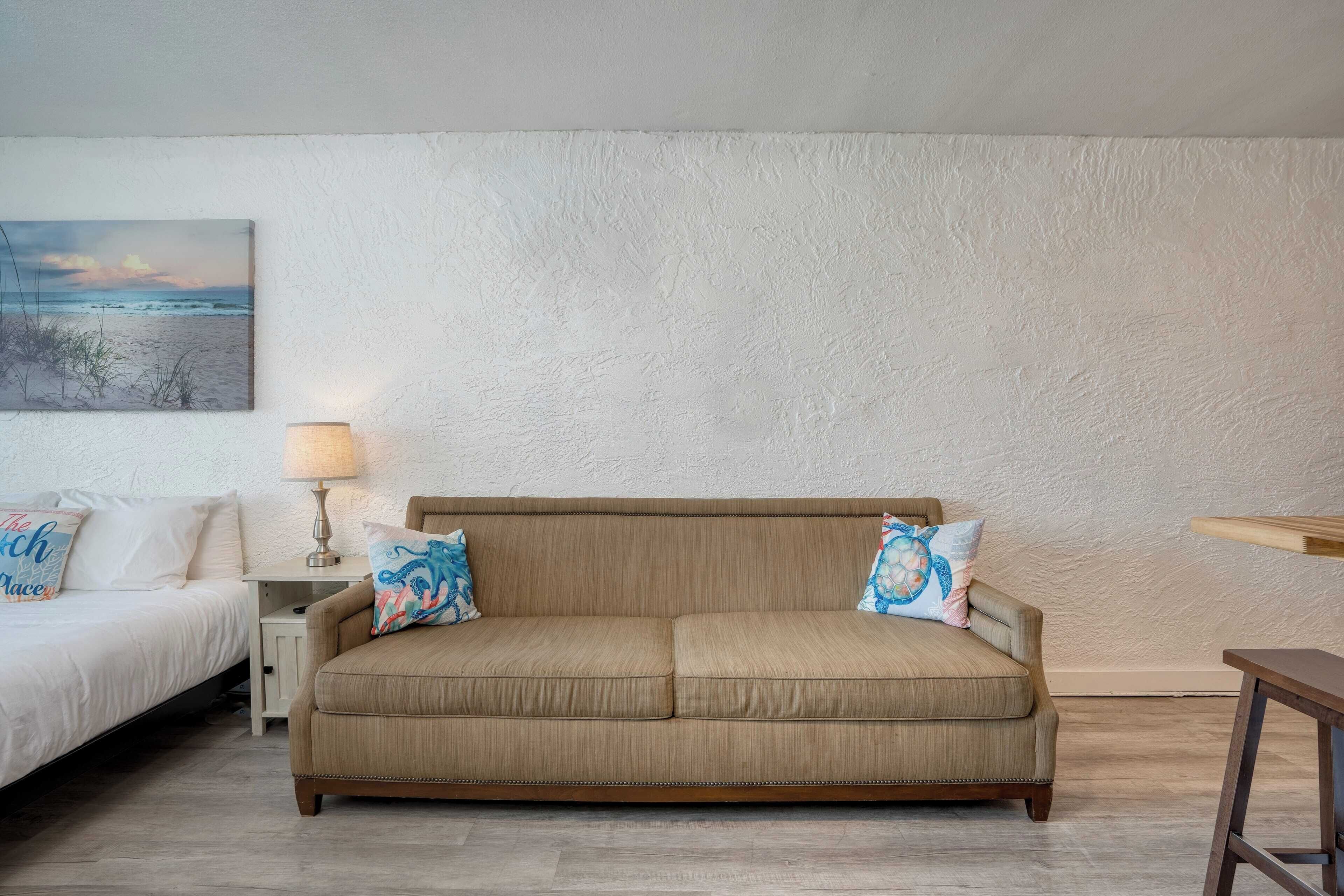 Una habitación con sofá color beige, una cama, una mesita de noche con lámpara y un cuadro que muestra una playa.