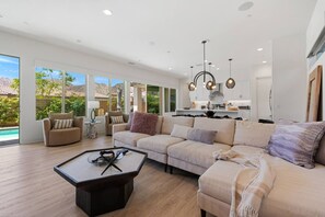 A spacious living room with a large sofa, a coffee table, and a view of a pool through large windows. - Casa Platino, Luxury PGA West home with Pool (La Quinta)